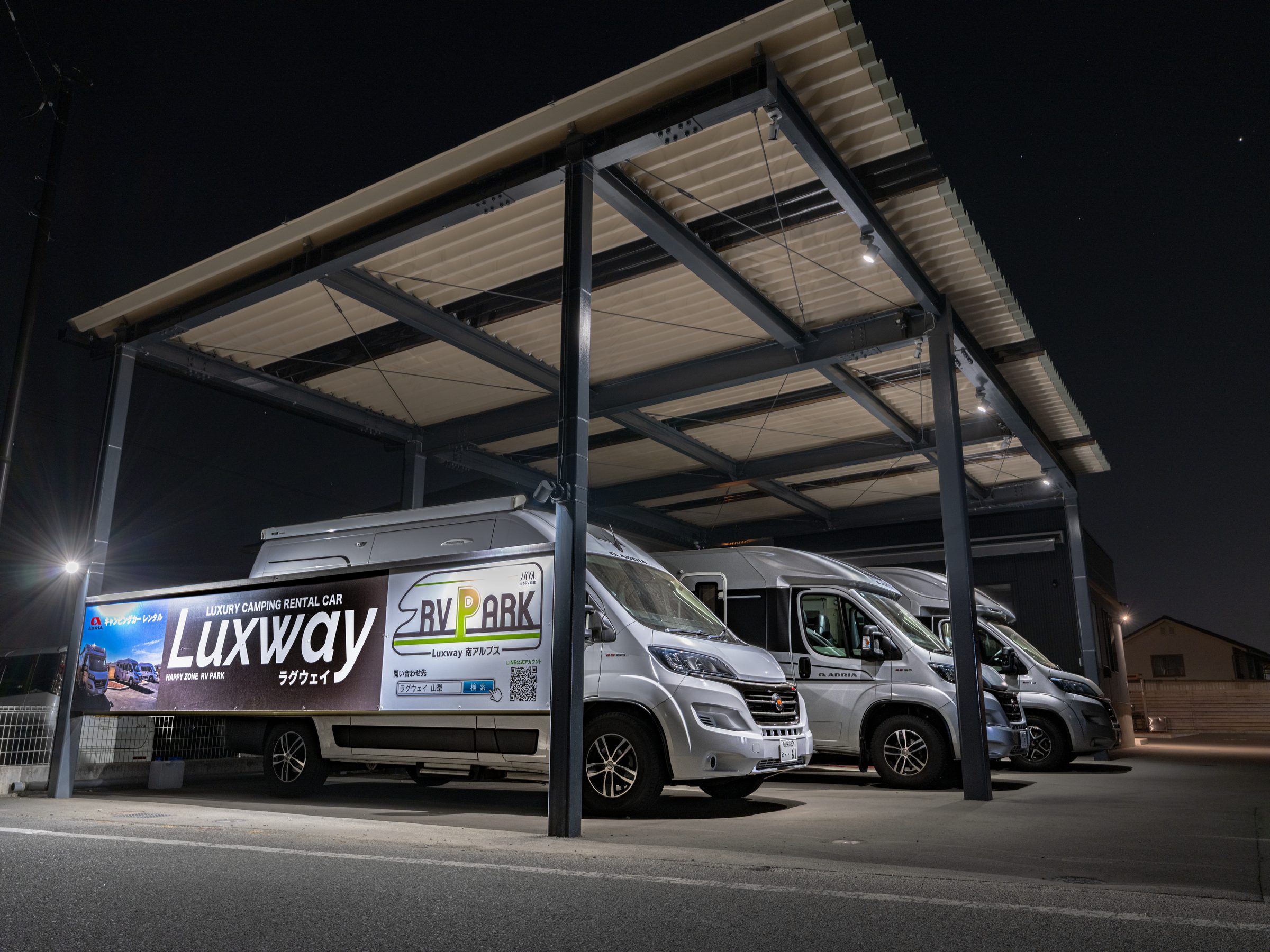 カーポートの下に並ぶ3台のADRIAのキャンピングカーと、Luxway／RV PARK 南アルプスの看板。夜