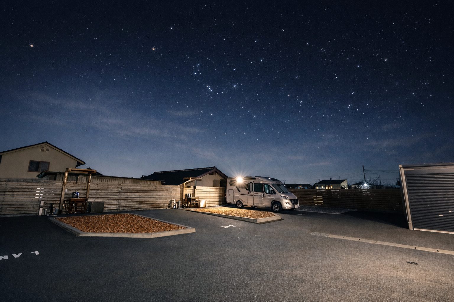 星空の下、瓦チップのフリースペースと木塀に囲まれたRVパークの区画。奥にキャンピングカーと管理棟の灯り