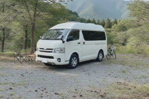 高原のキャンプ場の駐車スペースに停められた、無地で白いハイルーフ型バンコンバージョン・キャンピングカーを斜め後方から見た様子。背景に緑の木立と穏やかな山並み