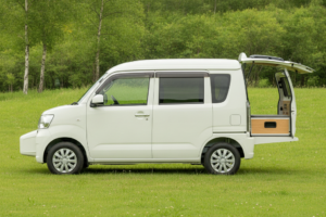 夏の高原のキャンプ場の芝生に停まる白い軽キャンピングカーを真横から見た様子。フロントウインドウの前に短いボンネットが張り出したFFレイアウトの軽バンで、バックドアが跳ね上げられ車中泊仕様の室内が見える