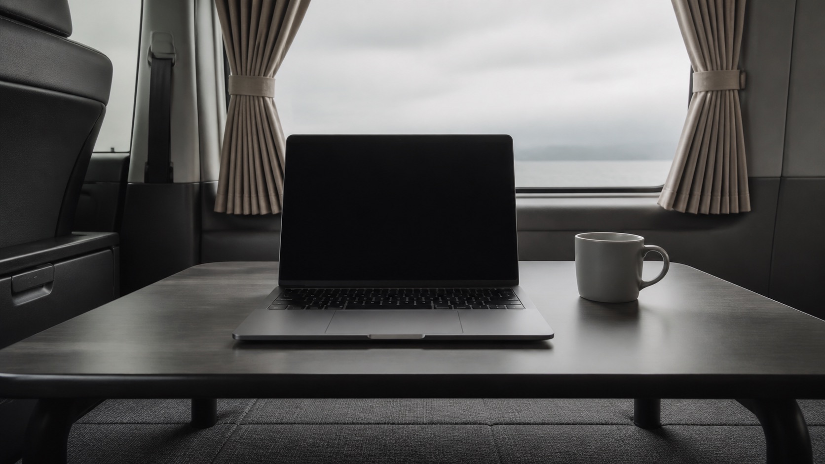 折りたたみの低いテーブルの上にノートPCとマグカップが並び、奥に車内の窓とカーテンが見える