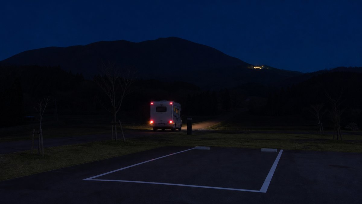 夜、RVパークの区画から本線へ出ていくキャンピングカーのテールランプと、遠くに見える温泉施設の明かり