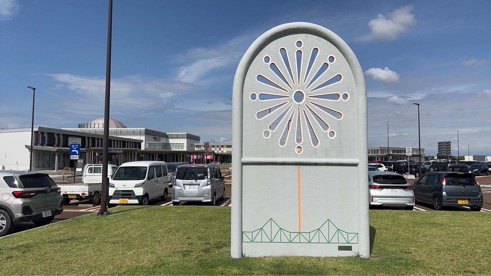 芝生に立つ花火モチーフの白いアーチ型モニュメント。背後に駐車場と建物