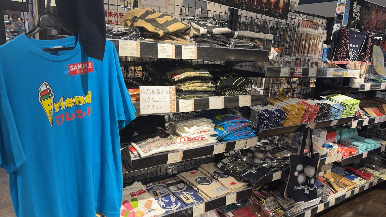 青いTシャツの見本品や手ぬぐい、花火柄のトートバッグが並ぶグッズ棚