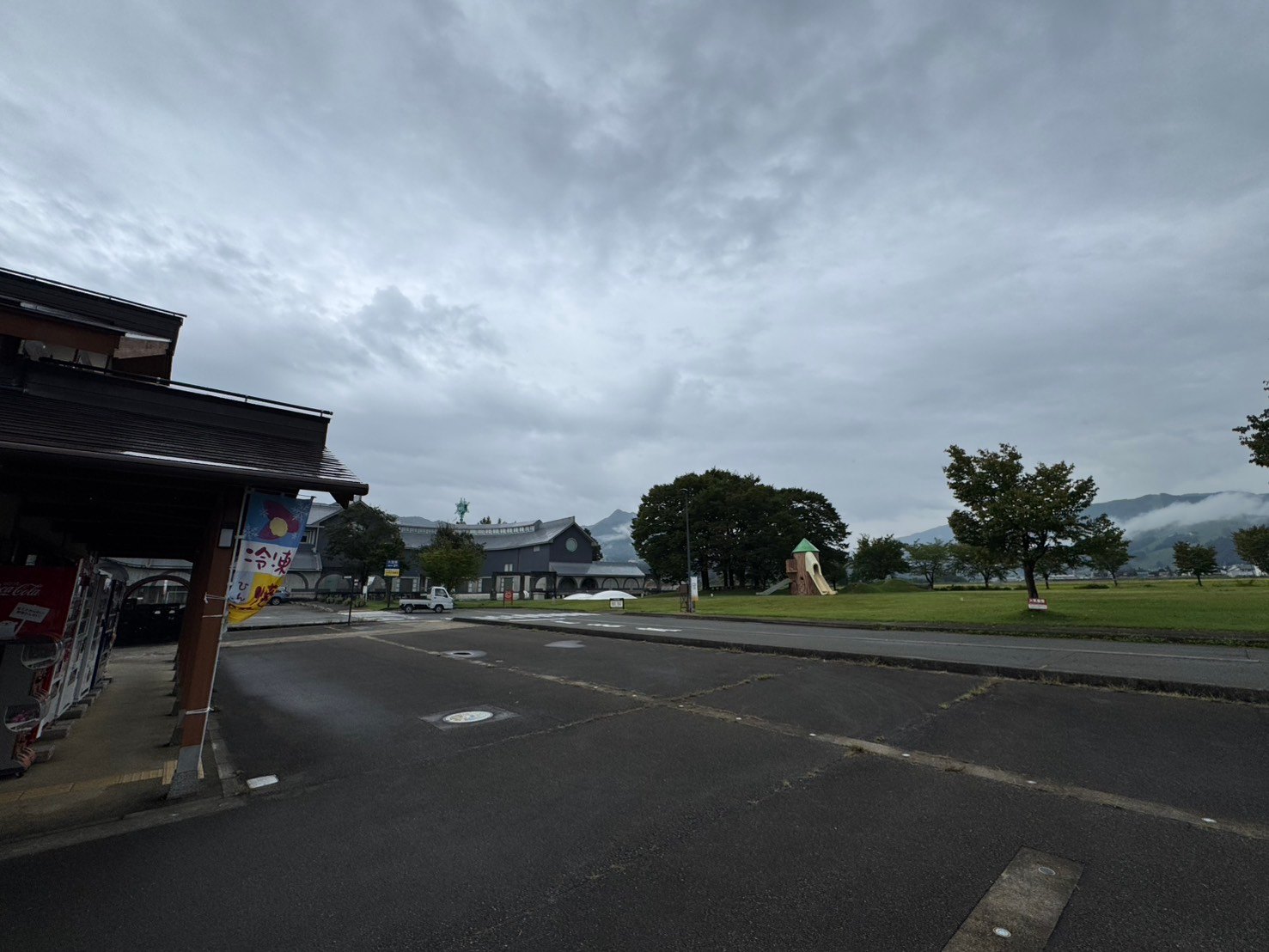 軒下から見た駐車場の先、道路の向こうに芝生の公園ととんがり屋根の滑り台遊具、木立と雲のかかる山並み