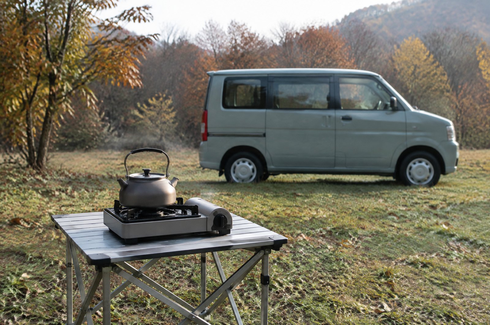 車から数メートル離して置いた折りたたみテーブルの上で、箱型のカセットこんろにかけたケトルで湯を沸かしている。車は奥にぼけて写っている。イメージ画像（AI生成。実際の商品の写真ではありません）