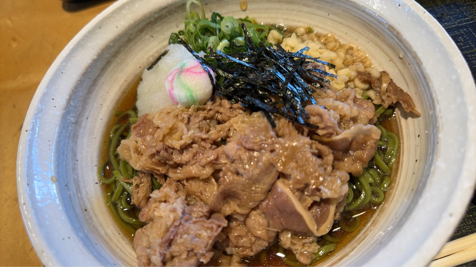 牛肉を広く盛った緑のそばに、刻みのり、ねぎ、大根おろし、丸い麩を添えた白い器