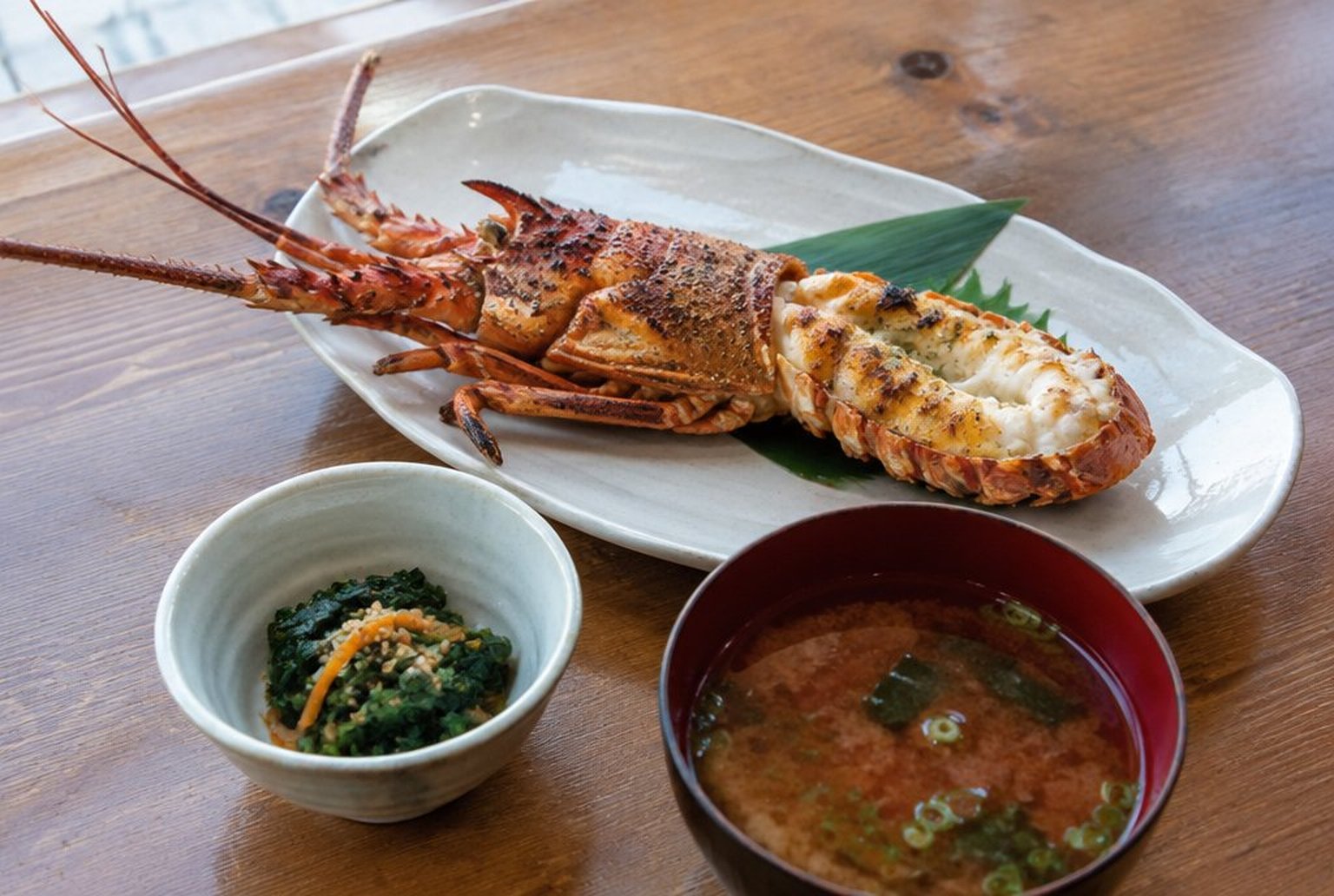 白い器に盛られた焼いた伊勢えびと、手前に小鉢と赤だしが並ぶ和食の御膳