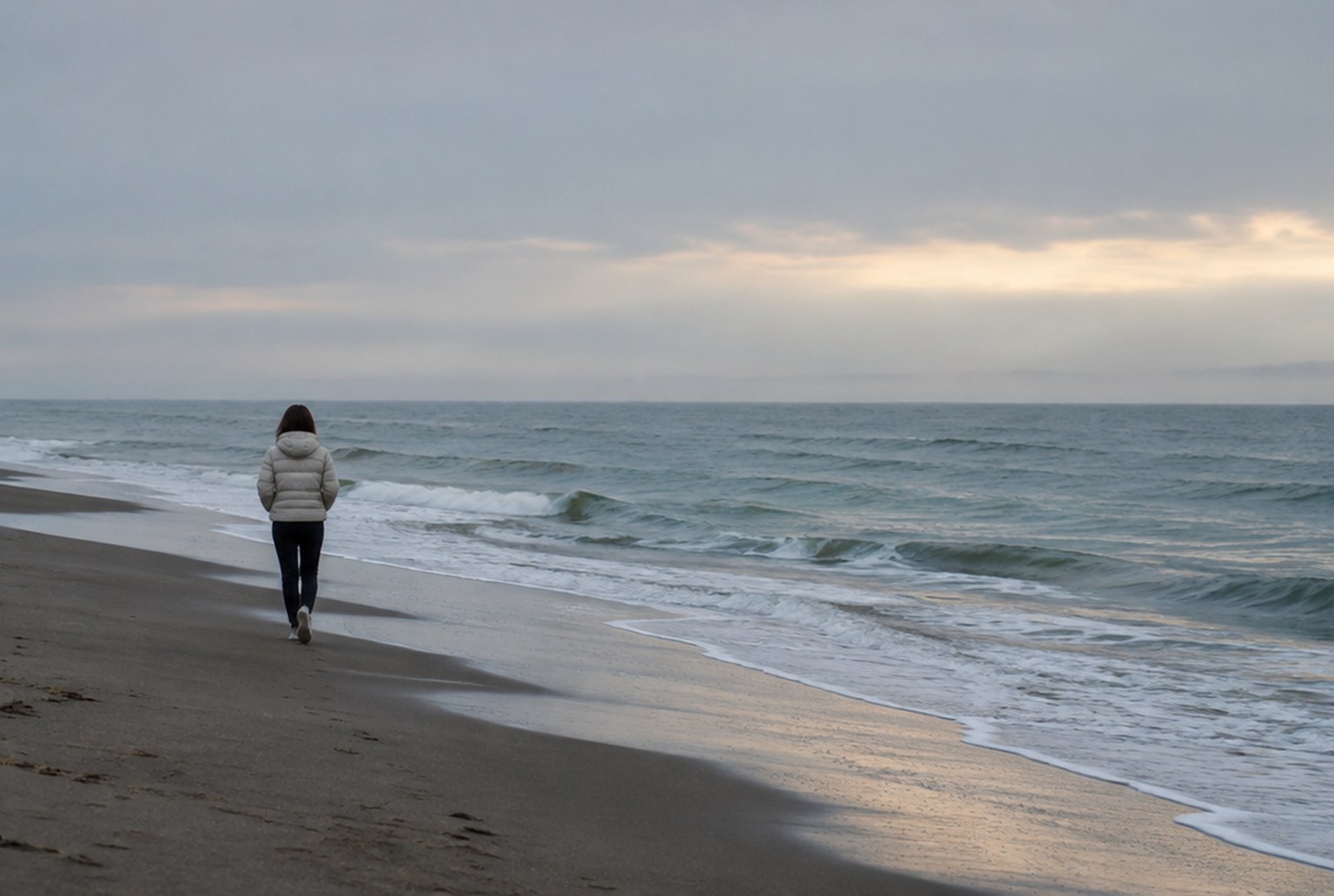 早朝、灰色がかった日本海の砂浜を、薄手のダウンを着た人がひとりで歩いている後ろ姿。波打ち際と薄い朝の光。建物・鳥居・看板はない。イメージ画像（AI生成。実際の施設・商品の写真ではありません）