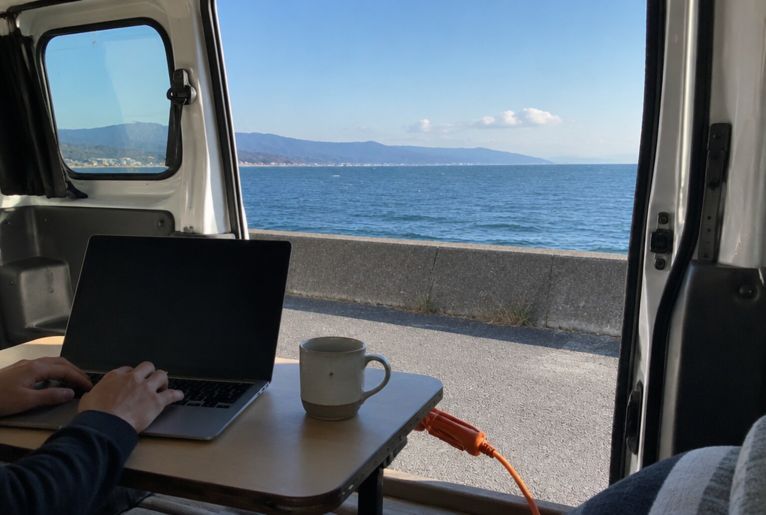 海沿いに停めたバンの車内。開けたスライドドアの向こうに日本海と防波堤、簡易テーブルにノートPCとマグカップ、キーボードに置いた手元。イメージ画像(AI生成。実際の施設・商品の写真ではありません)