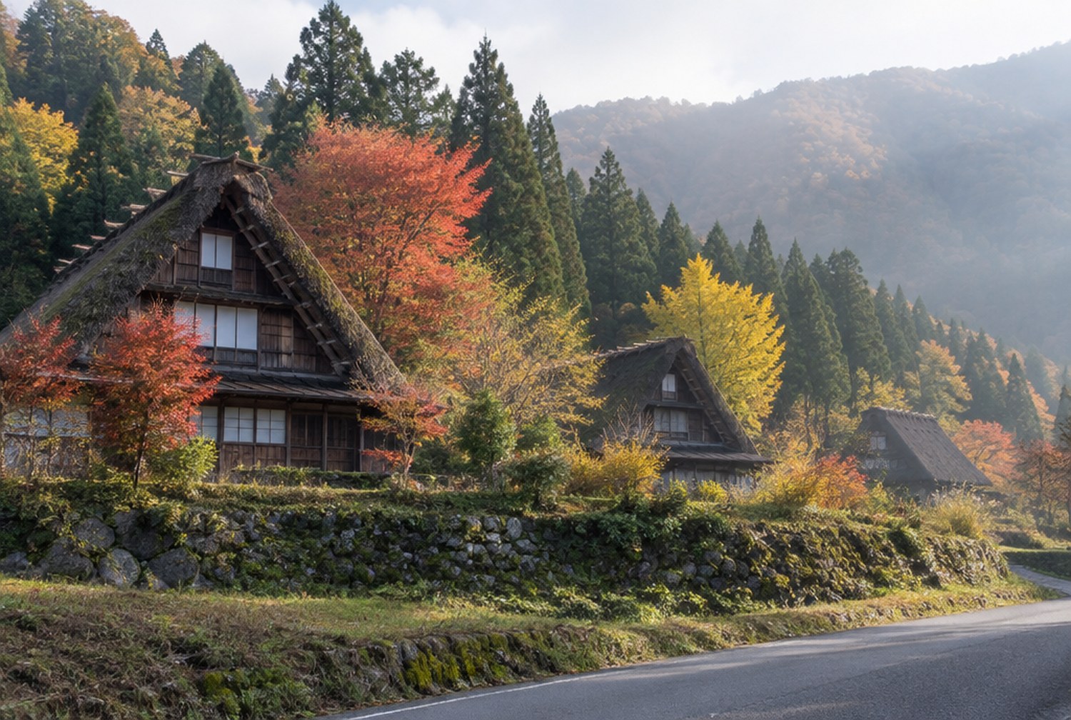 晩秋の山あいで、色づいた木々に囲まれた茅葺き屋根の古い民家が数棟並ぶ集落を、少し離れた道から見た風景。朝のやわらかな光。イメージ画像(AI生成。実際の施設・商品の写真ではありません)