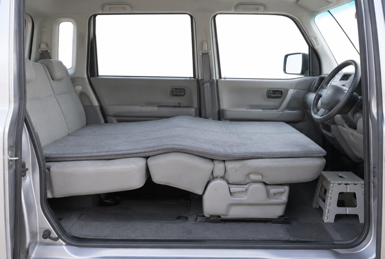 軽自動車の車内を開いたスライドドアの横から見た様子。前席の背もたれが後ろへ倒れて後席の座面とつながり、その上に厚手のマットが敷かれている。真ん中がわずかに盛り上がり、助手席の足元に小さな折りたたみ踏み台が置かれている(イメージ画像(AI生成。実際の施設・商品の写真ではありません))